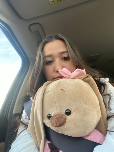 A young woman with long brown hair is sitting in the passenger seat of a car, holding a stuffed animal. She is looking directly at the camera with a neutral expression. The stuffed animal is a light brown bunny with black eyes and a pink bow on its head. The woman is wearing a white shirt. The car's interior is visible in the background, including the car's ceiling and a portion of the driver's side window. The sky and clouds are visible through the window. The image appears to be a selfie taken in the daytime. There is no information available about the location being in Staromusino, Russia.