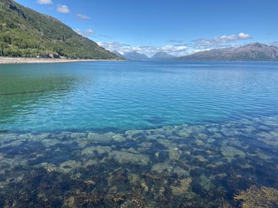 This media file depicts a scenic seascape in Lødingen, Norway, under a clear blue sky with scattered white clouds. The foreground features shallow, crystal-clear turquoise water revealing a rocky seabed with visible seaweed. The water transitions to a deeper blue further out.
The middle ground is dominated by a wide expanse of calm, blue water. In the background, a chain of majestic mountains, some with snow-capped peaks, stretches across the horizon. A road with a white campervan is visible running along the base of a green, tree-covered mountain to the left of the frame. This mountain is dotted with utility poles and wires. The overall scene evokes a sense of tranquility and natural beauty.
The time of day appears to be midday, given the bright sunlight and clear sky. The weather is fair and pleasant. The presence of mountains and the distinct coastline strongly suggest a Norwegian fjord or coastal landscape. No text is visible in the image.