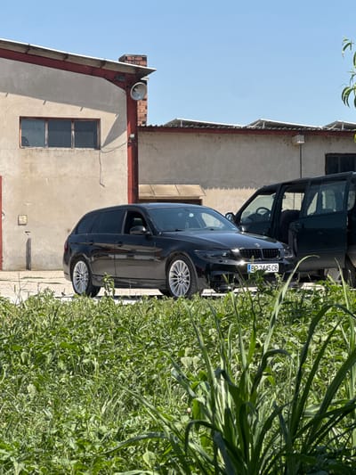 A black BMW touring car and a dark-colored van are parked in front of a light-colored industrial building with a red trim. The BMW's license plate reads "BP 2445 CB." The van's front passenger door is open. Tall green grass grows in the foreground, obscuring the lower portion of the vehicles. The scene appears to be outdoors under a clear blue sky. The location is identified as Belene, Bulgaria.