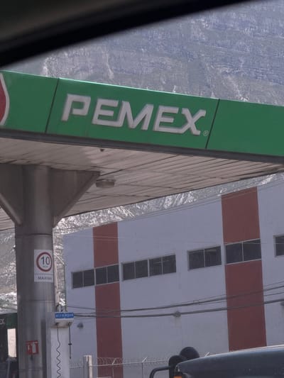 The image presents a static scene from a PEMEX gas station in Ciudad Santa Catarina, Mexico, viewed from within a vehicle, with the top and bottom edges partially obscured by the vehicle's interior. A large green canopy with the prominent white "PEMEX" logo dominates the upper portion of the frame, supported by a grey, cylindrical column. Attached to this column is a white circular sign displaying "10 km/h" and "MAXIMA" in black text. Below, an "aire agua" (air water) station is visible with hoses and a red fire extinguisher symbol. In the midground, a two-story building features a white facade with two wide vertical red stripes and multiple horizontal rows of dark rectangular windows. Utility lines traverse the scene in front of this building, and a chain-link fence topped with barbed wire is visible at its base. The background is dominated by large, grey, and partially vegetated mountains. No individuals are visible in the scene.
