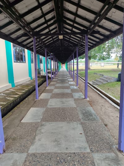 The image shows a long, covered walkway with a tiled floor. The walkway is bordered by a building on the left and a grassy field on the right. The building has white walls with turquoise accents and windows. The walkway is supported by purple pillars and has a dark-colored roof structure. The floor is a checkerboard pattern of large, light gray concrete tiles and smaller, darker tiles composed of pebbles embedded in concrete.
In the distance, the walkway extends towards a field where there appear to be targets, suggesting an archery or shooting range. A single person is visible walking on the field in the far distance. A large black trash bag is also visible on the grass near some concrete steps.
The scene appears to be outdoors, likely during the daytime, with natural light illuminating the walkway and the surrounding area. The presence of trees and greenery on the right side indicates a location with vegetation. The walkway creates a strong sense of perspective, drawing the viewer's eye towards the vanishing point in the distance. The overall mood is one of quiet observation, with no specific event taking place. The location is identified as Cotgadong, Indonesia.