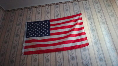 An American flag is draped on a wall. The flag is partially unfurled, showing its red and white stripes and the blue field with white stars. The wall behind the flag has a floral patterned wallpaper. The lighting in the image suggests an indoor setting, possibly a room. The location in the city of Barysh, Russia is not discernible from the visual information provided in the image.