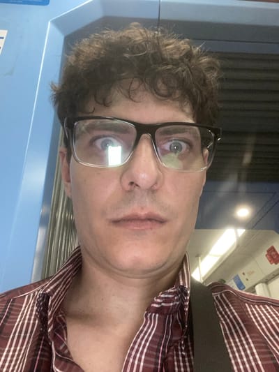 An adult male is depicted in a close-up, frontal self-portrait within a public transport vehicle in Luis Guillón, Argentina. The subject has dark, curly hair and is wearing black-framed glasses, through which his wide-open eyes with visible pupils are staring directly forward. Bright, rectangular light reflections are prominent in both lenses of his glasses. He is dressed in a collared, button-up shirt with maroon and white vertical stripes. A dark strap, consistent with a bag, is visible over his right shoulder. The environment suggests the interior of a train or subway car, featuring a light blue wall panel on the left with a partially visible white sticker, and a light-colored ceiling equipped with linear and circular overhead light fixtures. Dark horizontal structural elements are partially visible in the upper right background. No specific external landmarks or station identifiers are visible.