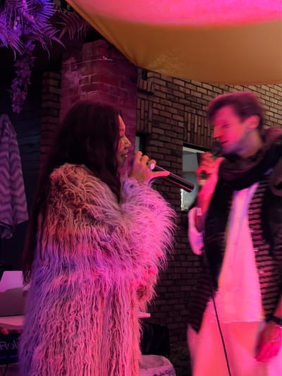 Two individuals are standing in front of a brick wall, illuminated by pink and yellow lighting. The person on the left, dressed in a long, shaggy pink coat, is holding a microphone and appears to be singing or speaking. Their face is partially obscured by their long, dark hair. The person on the right, wearing a dark top and a white garment underneath, is also holding a microphone and looking towards the left. They have short, light-colored hair and are positioned to the right of the first person. In the background, there are some artificial plants with purple lighting and a yellow awning. A keyboard with the brand name "Roland" is partially visible on the left side of the image. The scene suggests an outdoor performance or gathering, possibly at a venue in the city of Shebanovo, Russia.