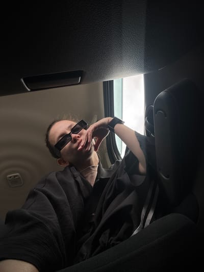 A person wearing sunglasses is angled towards the viewer, partially obscured by their hand. They appear to be inside a vehicle, possibly a car, with a sunroof visible in the background. The car's interior features dark upholstery and paneling. The location is unspecified, and there is no indication of the city of Bol'shoi Mashlyak, Russia.