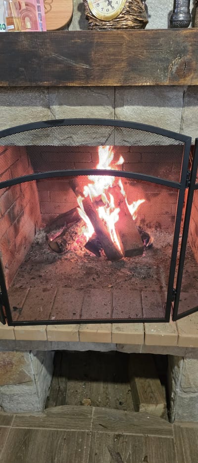The image shows a fireplace with a protective screen in Novachene, Bulgaria.  Burning logs are visible behind the screen. To the left of the fireplace, against a stone wall, there is a decorative wall clock, a wooden cutting board and some euro banknotes. To the right of the fireplace is what appears to be a niche in the wall. The construction material looks to be bricks.