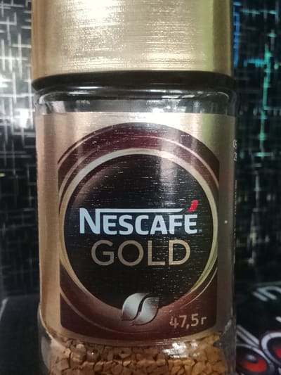 A close-up, slightly angled, eye-level shot shows the front of a glass jar of Nescafé Gold instant coffee. The jar is mostly filled with granular coffee. The label is predominantly gold and brown with a dark brown circular emblem in the center. Inside the emblem, in white, are the words "NESCAFÉ" and "GOLD". To the right of the emblem, the text "47,5r" is visible in white. The lid of the jar is a metallic gold color. The background is out of focus and appears to be a textured surface with a grid pattern and some blue and green lighting. There are no people or animals present, and no identifiable landmarks in the city of Obizarang, Uzbekistan are visible.