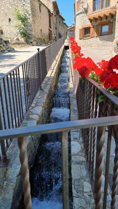 The image depicts a narrow, stone-lined channel carrying water, situated in the village of Setcases, Spain. The channel is bordered by metal railings on both sides. On the right side, a row of bright red flowers, likely geraniums, is planted in planters along the railing. The water in the channel appears to be flowing, with small cascades creating white foam. The surrounding architecture is composed of stone buildings with traditional wooden shutters and balconies. The scene suggests a picturesque, possibly historic, European village setting under a clear blue sky.