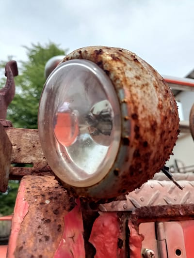 The image shows a close-up of a rusty, circular headlight on a weathered red vehicle. The headlight's glass is clear, revealing the bulb and a red reflection inside. The metal casing is heavily corroded with patches of orange and brown rust, and small bolts are visible around its edge. The headlight is mounted on a rusty metal bracket. In the background, out of focus, are green trees and a light-colored sky. The overall impression is one of age and disuse. The location is stated to be Blangbladeh, Indonesia, but there is no visual information in the image to confirm this specific location.