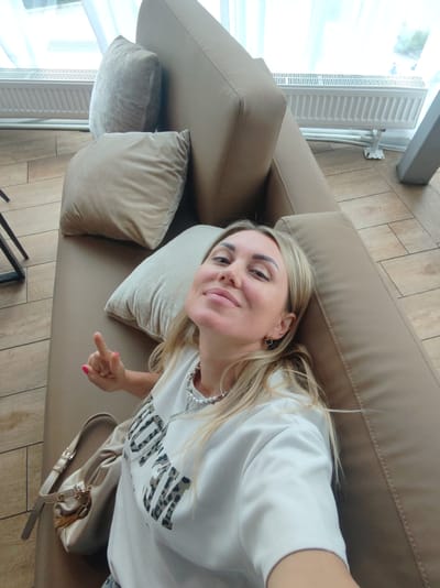 A woman with blonde hair is lying down on a beige sofa, looking up at the camera and smiling. She is wearing a white sweatshirt with a black and brown leopard print design that has text on it. She has a silver necklace with multiple strands and a small black earring in the shape of a clover. A beige purse with a gold clasp and tassels is resting on the sofa next to her. Her right hand is raised, with her index finger pointing upwards. Behind her, there are two light beige pillows and the armrest of the sofa. In the background, a white radiator is visible against a window with sheer white curtains. The floor is made of light brown tiles. The scene appears to be indoors, likely in a home or a public waiting area. The lighting suggests it is daytime, with natural light coming from the window. The woman's expression conveys happiness and playfulness. The text on her sweatshirt is partially visible and appears to be in a bold, block font.