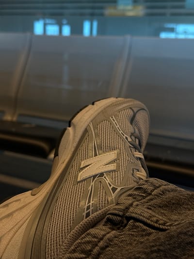 The image displays a close-up, angled view of a person's foot and lower leg, featuring a grey and white New Balance sneaker. The foot is positioned with the side of the shoe prominently visible. The shoe is primarily light grey with white accents, including the distinctive "N" logo on the side. It has a mesh upper, visible laces, and a multi-layered sole.
Part of the person's leg is covered by a dark grey, textured fabric, likely jeans or similar trousers, which appears slightly wrinkled.
The background is out of focus, showing blurry rectangular shapes and light reflections that suggest an indoor setting with windows or a modern architectural environment. The lighting is diffused, indicating either an overcast day or an interior space with ambient light. No specific activity is depicted; the foot appears to be resting.
The only visible text is the "N" logo on the side of the New Balance sneaker. The scene takes place in Weeze, Germany.