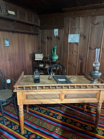 The image depicts an interior scene, likely a historical room within the city of Zheravna, Bulgaria. The room is paneled with dark wood, featuring shelves mounted on the walls. On the shelves, several books are visible, along with a framed picture and a decorative green pitcher.
The central focus is a wooden desk with intricate carvings and decorative balusters along the top edge. On the desk, there's an open ledger or notebook with a pen, an inkwell, a small object that appears to be a paperweight, and an antique oil lamp with a glass chimney. To the left of the desk, a chair with a woven seat is positioned, and a small white object, possibly a decorative item or a sensor, is attached to a string near the chair.
The floor is covered with a vibrantly colored rug featuring a geometric pattern of stripes in red, blue, yellow, green, and black. The overall impression is of a preserved historical study or office, evocative of a past era.