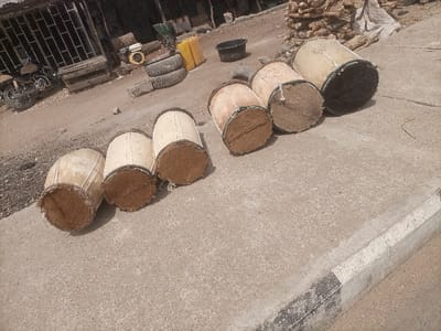 The scene is an outdoor commercial or roadside area in Gulum, Nigeria. Six light-colored, cylindrical drums, appearing to be crafted from wood or similar natural material with fibrous ends, are arranged horizontally on a concrete pavement. The drum on the far right displays a dark, possibly animal hide, surface at one end. A concrete curb with alternating black and white paint delineates the pavement from an unpaved dirt area. Within this dirt area, a dark motorcycle is parked on the left. Further right, stacks of tires, a yellow portable generator, several yellow plastic jerrycans, and a black basin are visible. A large pile of stacked firewood or timber occupies the upper right background. Commercial structures with metal security grates are present in the far left background. No individuals are depicted, and no specific actions or interactions are occurring.