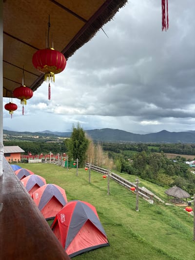 The scene depicts an outdoor elevated perspective overlooking a valley in Ban Mai Saha Sampan, Thailand, under an overcast sky. In the immediate foreground, the underside of a light-colored thatched roof structure is visible, from which three red Chinese lanterns with gold accents and red tassels are suspended. A fourth red lantern is partially visible to the left. A dark wooden railing angles across the bottom left of the frame.
The midground features a descending grassy hillside. Four red and grey/blue dome-shaped camping tents are pitched sequentially on the grass, appearing unoccupied. To the right of the tents, a series of vertically oriented wooden posts with crossbars lines the slope, each supporting a smaller red Chinese lantern. A wooden fence structure runs parallel to these posts further down the hill. A circular, open-sided gazebo with a dark thatched roof stands further down the slope to the far right.
In the background, a vast landscape extends, characterized by rolling hills covered in dense green vegetation and forested areas. Scattered buildings with orange or red roofs are discernible amidst the greenery. Distant mountains form the horizon line under a heavily clouded, grey sky, indicating potential rain. No individuals are visible.