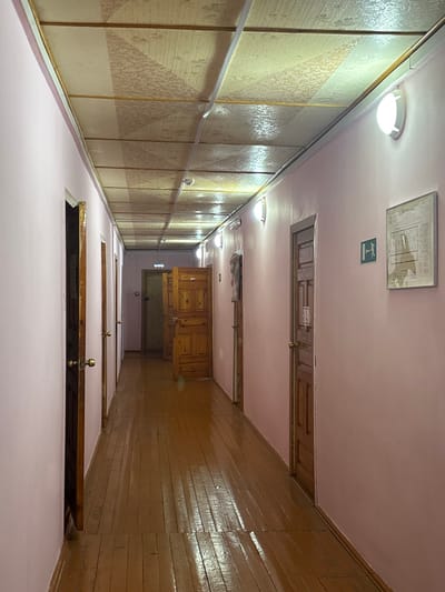 This image presents an interior view of a long, narrow hallway located in Bayronovka, Russia. The hallway features light pink walls and a dark brown, varnished wooden floor exhibiting reflections from overhead light sources. The ceiling consists of light-colored, textured panels arranged in a grid with visible support structures. Illumination is provided by multiple circular, wall-mounted light fixtures with white diffusers, evenly spaced along both walls, along with subtle overhead lighting. Numerous wooden paneled doors, equipped with brass-toned doorknobs, line both sides of the hallway; some are closed, while others are slightly ajar. Towards the far end of the hallway, an open doorway reveals an interior space, and a sign above it appears to indicate an exit. On the right wall, a green rectangular emergency exit sign with a white running-man pictogram and a right-pointing arrow is affixed, adjacent to a framed document, possibly an emergency plan or informational notice. The hallway is unoccupied.