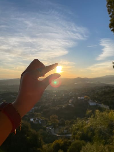 The image, presented in a landscape orientation, captures a human hand seemingly "pinching" the setting or rising sun. The hand, positioned in the middle-right, is extended upwards with the index and middle fingers separated, framing the bright golden sun between them. A silver-toned ring is visible on the middle finger, and a red or maroon wristband adorns the wrist.
The sky occupies the left half of the frame, transitioning from a clear blue at the top-left to a radiant golden hue around the sun, illuminated by the low-lying light. Delicate, wispy white clouds streak across the blue expanse. A distinct green lens flare is noticeable to the right of the hand, resulting from the direct sunlight hitting the camera lens.
The right half of the image showcases a sprawling, verdant landscape stretching into the distance. This hilly or mountainous terrain is covered in dense green foliage, with faint hints of winding roads or structures nestled within, suggesting human presence in the natural environment. The warm, soft light indicates the golden hour, either sunrise or sunset, casting long shadows and a gentle glow over the scene. The overall ambiance is one of serenity and natural beauty, characteristic of a scenic overlook in Mijas, Spain. No discernible text is present in the image.