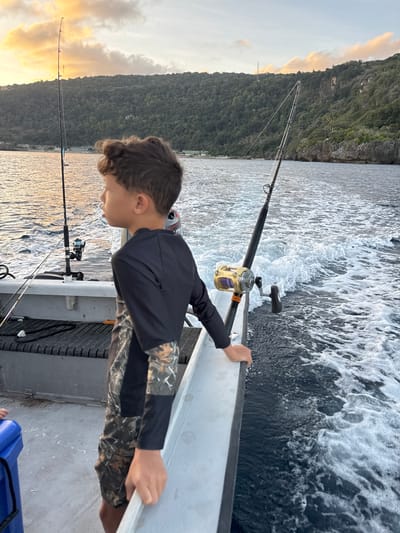A young boy with dark curly hair, wearing a long-sleeved black shirt with camouflage sleeves and matching camouflage shorts, stands on the deck of a boat. He is looking out to the left of the frame, with his right hand resting on the boat's railing. Two fishing rods are visible, one with a gold reel and purple fishing line, and another with a silver reel. The boat is moving through dark blue water, creating a significant wake. In the background, a lush green, tree-covered island with rocky cliffs rises from the sea. The sky above is partly cloudy with hues of orange and yellow, suggesting either sunrise or sunset. The location is the waters surrounding Christmas Island, Australia.