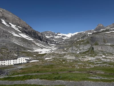 The image shows a wide mountain landscape under a clear blue sky. In the foreground, a large, white, multi-story building complex with numerous windows is situated on a grassy slope. The building appears to be an older hotel or lodge. To the left of the building, there is a pile of dark rubble.
The middle ground is characterized by rolling, rocky terrain interspersed with patches of green grass and snow. A winding road is visible in the distance, leading up the mountainside. A tall metal pylon for power lines is also present near the building complex.
The background is dominated by imposing, rugged mountain peaks. Some of these peaks are snow-capped, and streaks of snow are visible on the rocky slopes. The scale of the mountains suggests a high-altitude environment. The overall impression is one of a remote, alpine setting. The scene is likely located in the vicinity of Leukerbad, Switzerland, known for its mountain scenery and thermal baths. There are no people visible in the image.