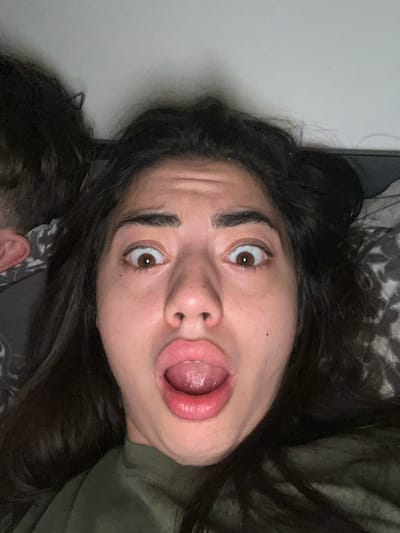 The image shows a close-up, overhead view of a young woman lying down. She has dark, wavy hair and is wearing a dark green shirt. Her eyes are wide open, and her mouth is open with her tongue slightly sticking out. The background appears to be a bed with a patterned comforter. There is no visible indication of the city of Chotěšov, Czechia.