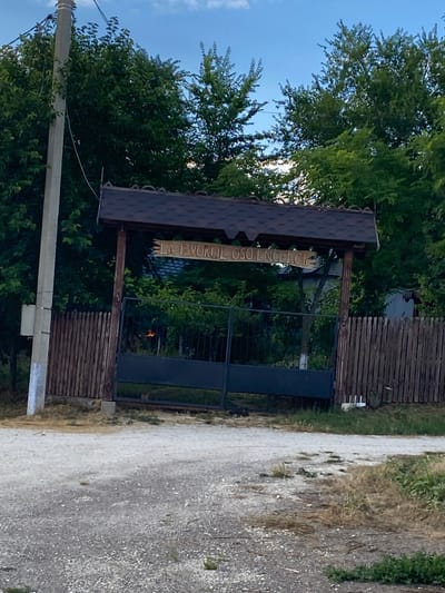The image shows an entrance gate to a property located in Horești, Moldova. The gate is made of dark metal and is flanked by a wooden fence. Above the gate is a wooden archway with a brown tiled roof, featuring a carved sign that reads "LA IZVORUL OSOENCELOR". A concrete utility pole stands to the left of the entrance, with wires extending from it. The surrounding environment consists of lush green trees and vegetation, with a gravel road leading up to the gate. The sky is a clear blue with some scattered clouds. The overall scene depicts a rural entrance to a property.