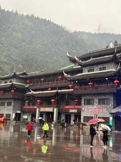 The image presents an outdoor scene in Pengshui Miao and Tujia Autonomous County, China, under conditions of rain or recent rainfall, indicated by the extensively wet and reflective paved ground. The central subject is a large, multi-story building featuring traditional Chinese architecture with dark gray brick walls, multiple tiers of dark tiled roofs with distinctive upturned eaves, and ornate wooden balconies or railings on each level. Numerous red Chinese lanterns are suspended along these balconies and below the eaves. Red banners with white Chinese characters are affixed to the building's lower facade; one banner prominently displays "游客中心" (Tourist Center).
In the foreground, a wide, dark gray paved plaza is thoroughly soaked, creating strong reflections of the building and individuals. Approximately 10-15 people are visible on the plaza. On the left, a woman wearing a red jacket walks rightward. Near the center, a man in a bright yellow-green jacket walks in the same direction. On the right side, two individuals stand together, one holding a solid red umbrella and the other a light multi-colored umbrella. Several other figures are dispersed across the plaza, some holding umbrellas.
The background consists of a densely forested mountain or large hill, partially obscured by atmospheric haze or mist. A power transmission tower is faintly visible on the upper right slope of this distant hillside.