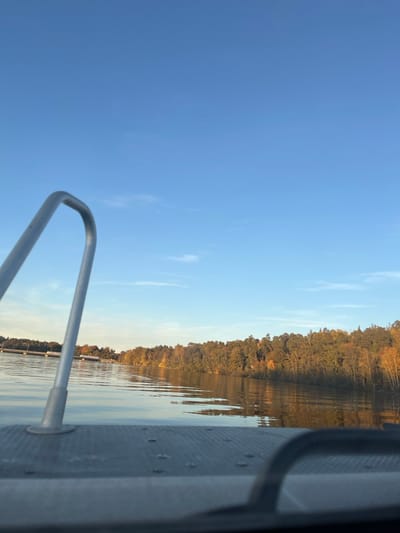The image depicts a tranquil outdoor water scene, observed from a low vantage point on a vessel within Ekerö kommun, Sweden, likely on Lake Mälaren. The upper portion of the frame features a clear, bright blue sky with sparse, wispy white clouds. The middle ground is occupied by a calm body of water, exhibiting minor ripples and reflections of the sky and distant land. Along the distant horizon, a shoreline is visible, densely covered with trees displaying prominent autumn foliage in shades of green, yellow, and orange. A light-colored, linear floating structure, consistent with a dock or pier, is visible on the far left of the distant shore. In the immediate foreground, a textured, light gray deck surface of a boat is visible, studded with circular fasteners. A curved, metallic railing or handhold extends from the lower-left, ascending towards the center of the frame. The scene is illuminated by natural sunlight, suggesting a clear day during autumn. No individuals are present.