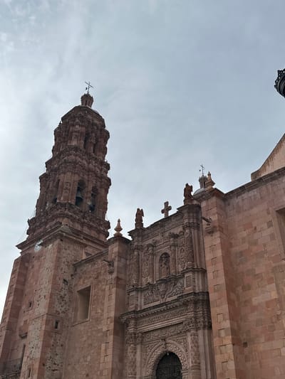 The image shows a low-angle view of the exterior of the Zacatecas Cathedral in Zacatecas City, Mexico, under an overcast sky. The cathedral is constructed of pinkish-red stone. A tall, ornate bell tower, also made of stone, dominates the left side of the frame. The tower features multiple levels with arched openings where bells are visible, intricate carvings, and a cross at its apex. A clock face is also visible on the lower part of the tower. To the right of the tower is the main facade of the cathedral, characterized by elaborate baroque carvings, niches with statues, and arched doorways. The building's facade is also made of the same reddish stone. The overall impression is of a grand and historically significant religious structure.