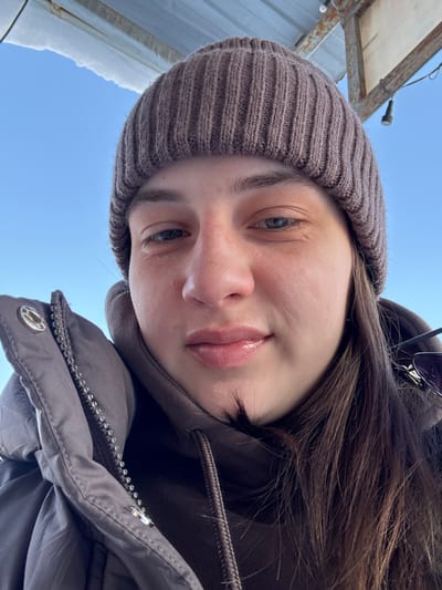 A selfie-style shot looking upwards at a fair-skinned person with brown hair and blue eyes. They wear a brown knitted beanie and a dark brown hooded jacket zipped to the chest. The background is a bright blue sky with a partial view of a metallic structure and snow. The subject's face is tilted, lips appearing full and glossy. The image is slightly overexposed, creating a soft, diffused light.
