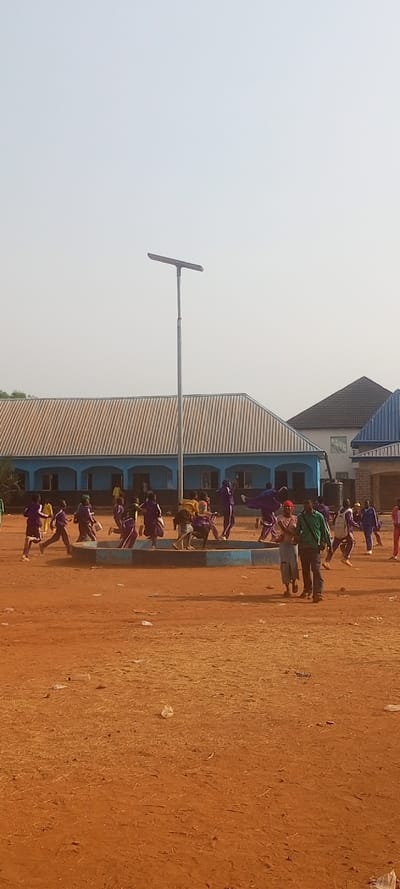 The image depicts an outdoor scene in Tuaniako, Nigeria, under an overcast or hazy sky. The foreground consists of reddish-brown dirt and dust, sparsely covered with dry vegetation and scattered litter, including white plastic bags.
A tall, grey metallic pole with a rectangular solar street light fixture at its top stands centrally in the midground. Surrounding the base of this pole is a low, blue and white painted octagonal or circular structure, possibly a planter or a boundary.
Behind this structure and pole, a single-story building with blue walls and a light-colored corrugated metal roof extends across the frame. This building features multiple arched doorways and windows. Further in the background, additional residential-style buildings with dark and blue roofs are visible.
Numerous individuals are present, predominantly children appearing to be in school uniforms of varying shades of purple, blue, and yellow. They are actively engaged in running and playing across the dusty ground, particularly around the blue and white structure. Their movements suggest dynamic activity. Two adults are observed in the mid-foreground, walking from right to left. One male wears a green long-sleeved shirt and dark trousers. The other adult, a female, wears a light-colored patterned top and a light skirt, possibly holding hands with the male. All individuals are oriented towards the activities or moving through the space.