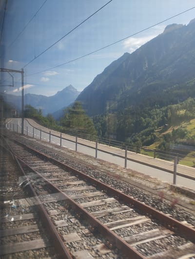 The image depicts a view from a moving train, looking out at a mountainous landscape. In the foreground, railroad tracks stretch into the distance, with a road running parallel to them, separated by a metal guardrail. The road curves around a hillside covered in lush green trees. Beyond the road, the landscape rises dramatically into steep, verdant mountains, with a clear blue sky above. The sun casts a warm glow on the scene, highlighting the textures of the foliage and the ruggedness of the mountains. In the middle ground, a small, isolated building can be seen nestled on a grassy slope. The overall impression is one of natural beauty and tranquility, with the man-made elements of the train tracks and road integrated into the environment. The image is likely taken in the vicinity of Wassen, Switzerland, a region known for its stunning alpine scenery and extensive railway network.