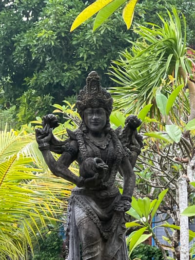 A gray statue of a woman with multiple arms stands amid lush greenery. She wears an ornate crown and a decorative necklace. Her face is serene, and her hands hold various objects. Palm fronds and other tropical foliage surround the statue, creating a vibrant, natural backdrop. Some leaves appear yellow, while the rest are green.
