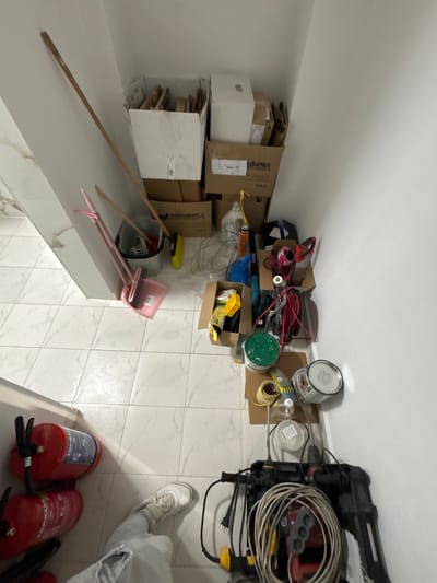 In the city of Madan, Bulgaria, a corner of a room is shown, likely a storage area or a workspace. The floor is tiled with a white, square pattern. Against the white wall, various items are stacked and leaning.

To the left, a bright red fire extinguisher is visible, mounted on the wall, with another one below it. Further to the left, part of a person's leg wearing ripped light-colored jeans and a white sneaker is visible, suggesting someone is standing in or near the frame.

The main area is filled with a collection of household and construction-related items. At the top left, a long wooden pole, possibly a broom handle, leans against the wall, next to a large white cardboard box. Below this, a pink dustpan and brush set is propped up. To the right of the dustpan, a grey bucket contains what appears to be cleaning tools. Further right, several cardboard boxes are stacked. One box has Cyrillic lettering that appears to say "ЧАЙКАФАРМА" (ChaikaFarma), indicating it's likely a pharmaceutical or healthcare product box.

The items become denser towards the right side of the image. There are rolls of tape, various containers, including a green-lidded one and a silver-colored can. Electrical cords are coiled and strewn about, and a power tool, possibly a drill, is visible at the bottom right, with a coiled grey and yellow cable connected to it.

The lighting in the room is bright, casting some shadows, and the overall impression is one of a somewhat cluttered but functional storage space.