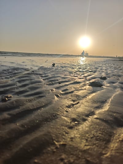 The image depicts a low-angle view of a beach at sunset. The foreground is dominated by the textured, rippled sand left by receding tides, with small pebbles scattered across it. The wet sand reflects the golden light of the setting sun, creating a shimmering effect. In the distance, the sun is a bright orb low on the horizon, casting long rays of light across the sky and water. Silhouetted against the sun are faint structures, possibly industrial buildings or port facilities, suggesting an urban or industrial coastal setting. A solitary figure, also in silhouette, walks towards the sun on the wet sand. The sky is a soft gradient of pale blue to warm yellow, indicative of sunset. The overall mood is serene and tranquil, capturing the peacefulness of a beach at the end of the day. The location is identified as El Puerto de Santa María, Spain.
