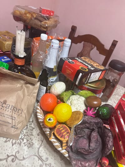 The image shows a cluttered tabletop scene. Prominent items include: a box of "Panecillos Premium" cookies, various bottles (some labeled "Tinelli" and "Reposado"), a brown paper bag from "Ccatori Pizzavino, Miguel Hidalgo #2, San Cristóbal de las Casas, Mexico," a basket containing an assortment of artificial fruits and vegetables, and miscellaneous packaged goods. A partially burned white candle is visible near the bottom of the frame.