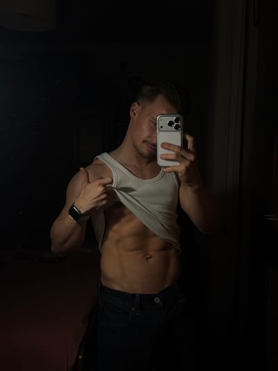 A person stands in dim lighting, holding a phone to take a selfie. The individual wears jeans and a white tank top, partially lifted to reveal abdominal muscles. A watch is visible on the left wrist. The background is dark and indistinct, possibly a bedroom or living space. The image appears to be a self-portrait intended to showcase the person's physique.