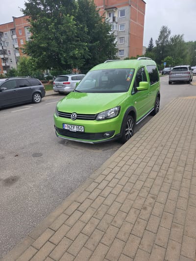 A bright green Volkswagen Caddy is parked on the right side of a two-lane asphalt road in Širvintos, Lithuania. The vehicle is facing forward, slightly angled towards the right edge of the frame. Its license plate reads "NZF 150". To the right of the car, a sidewalk paved with tan rectangular bricks runs parallel to the road, separated by a concrete curb. Behind the Volkswagen Caddy, a silver Volkswagen Passat wagon is parked further down the road. In the background, a multi-story apartment building with a reddish-orange facade and grey accents stands behind a cluster of lush green trees. The sky is overcast and appears to be a light grey.