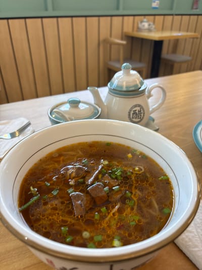 The image depicts a close-up view of a bowl of noodle soup, likely beef noodle soup, on a wooden table in a restaurant. The soup is a rich brown color with visible pieces of meat and chopped green onions. The bowl has a white exterior with a black rim and a decorative floral pattern at the bottom. To the left of the soup bowl, a fork is placed on a white napkin. In the background, a small white teapot with blue accents and a lid sits on the table, next to a lidded condiment dish. The restaurant interior features light-colored vertical wooden paneling on the walls. In the further background, a table with chairs is visible. The image's lighting suggests an indoor setting, possibly during daytime. The exact location within Putrajaya, Malaysia, cannot be determined from the visual information provided.