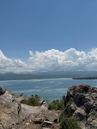 This is a vertical, eye-level, outdoor, wide-angle photograph taken from a rocky outcrop overlooking a large body of water. The scene is dominated by a vast expanse of calm, blue water that fills the right two-thirds of the frame. Distant, hazy mountains, some with patches of snow, form a backdrop to the water. On the left side of the image, a dramatic wall of white, puffy cumulus clouds rises from the horizon, contrasted against a clear, bright blue sky. The rocky foreground on the right is rough and textured, with sparse green vegetation, including some small yellow wildflowers, growing in the crevices.
The sun appears to be high in the sky, casting clear light and suggesting a warm, sunny day. The weather is fair, with large clouds indicating it might be a bit breezy. The overall mood is serene and majestic, capturing the natural beauty of a mountainous landscape and a large lake. There is a small, bright pink object visible in the far left of the sky, its nature unclear. No people or specific objects are discernible, but the scene clearly depicts a natural landscape, likely a lake such as Lake Sevan in Armenia, given the context provided. The time of day is likely midday or early afternoon due to the bright sunlight and cloud formation.