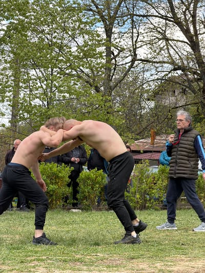 Two shirtless men are wrestling in a grassy field. Both are wearing black pants and black shoes. An older man wearing glasses, a dark puffer vest, and dark pants watches the wrestling match. Other people are also observing in the background. A tree with budding green leaves is in the background. The location is Dulevo, Bulgaria.