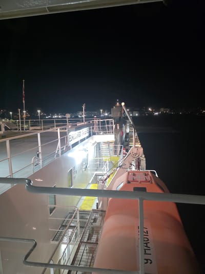 The image shows a nighttime view from the deck of a ferry in Brindisi, Italy. The ferry, identified by the text "EUROCARGO TRIESTE" on its side, is docked or moving slowly. The scene is illuminated by artificial lights from the ferry and the distant cityscape. A red life raft is visible on the right side of the ferry, with some text "Y HAAIZT" printed vertically on it. A flag, possibly Italian, is flying on a flagpole on the left side of the ferry. In the background, the lights of Brindisi are visible along the coastline, reflecting on the dark water. The environment is maritime, with the ferry dominating the foreground and the city lights in the distance suggesting its proximity to land. There are no people visible in the frame.