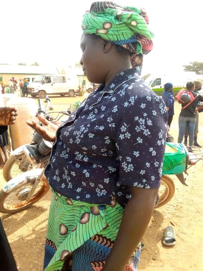 A woman wearing a blue, floral-patterned blouse and a colorful green and blue patterned wrap stands near a parked motorcycle. She also wears a multi-colored headwrap. People and vehicles are visible in the background near a building. A single sandal rests on the ground. The ground appears to be dirt or sandy soil.