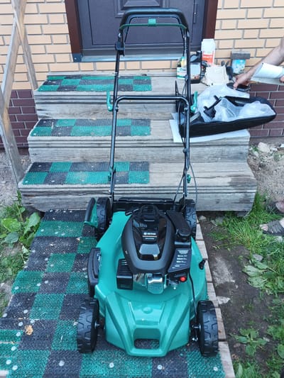 A medium-shot, outdoor, eye-level view shows a new, bright green lawnmower positioned on a black and green checkered mat laid out on wooden steps. The lawnmower has a black engine cover with a silver logo and some text, and a black handle with a green adjustment knob. A black bag, partially covered in plastic bubble wrap, sits on a wooden surface behind the lawnmower. Several bottles and packets are also on the wooden surface. A person's hand is visible holding a document, suggesting they are either reading instructions or showing them to someone. The background consists of a brick wall and a dark brown door, with some wooden planks and green grass visible on the ground. The scene appears to be in a residential outdoor setting, likely a yard or porch area, with a hint of a building under construction or renovation due to the presence of unpainted wooden planks. The lighting suggests it is daytime, possibly late afternoon, with soft light. No specific weather conditions are evident, but the grass is green and appears healthy. Visible text includes "БЕЛАЯ" and "BAX.07246" on the lawnmower's engine cover, and some unreadable text on the bottles. The overall mood is neutral, possibly indicating the assembly or preparation of the lawnmower for use.