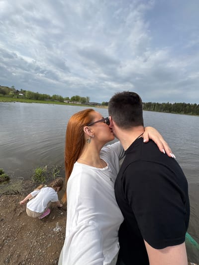 The image captures a couple kissing near a body of water in Nozhovka, Russia. The man wears a black shirt, while the woman has long, reddish-brown hair and a white shirt. A child in a white shirt and khaki pants kneels near the water's edge, seemingly examining the ground. The environment includes a lake, trees, and cloudy sky.