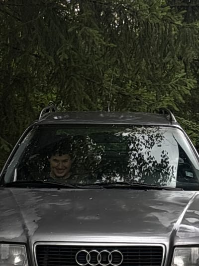 A man is visible through the windshield of a car, smiling broadly. He is sitting in the driver's seat. The car is dark gray and appears to be an Audi. The scene is outdoors, with dense green foliage behind the vehicle. It is unclear if the car is in Taban, Bulgaria, or any other city.