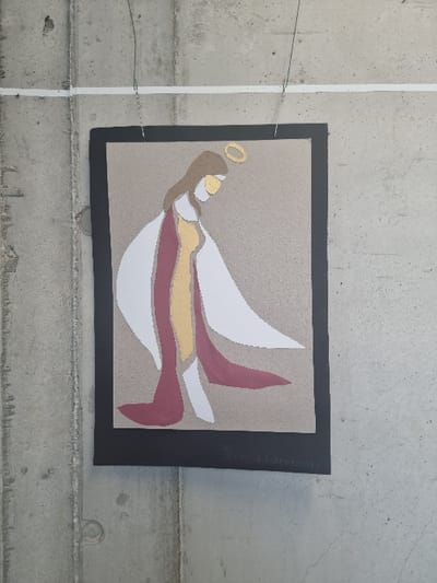 An abstract, stylized artwork depicting a figure resembling an angel with a golden halo is suspended against a grey concrete wall in Jaunmārupe, Latvia. The artwork, framed with a black border, features a central figure rendered in white, gold, and dark red shapes on a light brown background. The figure, shown in profile facing left with a bowed head, has brown hair and a light-colored face. The bottom right corner of the artwork bears a signature, "Māris Caunāns," and the year "2022." The piece is hung by two thin wires with hooks, which are attached to a narrow white horizontal strip positioned high on the wall. The concrete wall itself is unadorned, exhibiting typical texture and minor imperfections.
