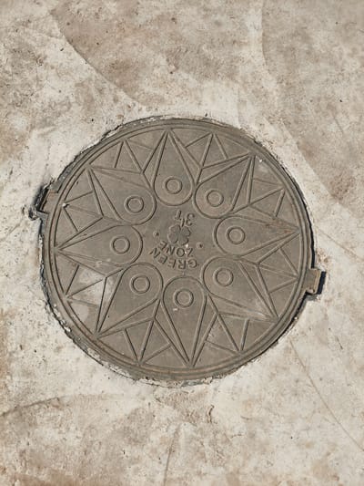 A circular metal manhole cover is embedded in a light-colored concrete surface. The manhole cover features an intricate starburst pattern with triangular and circular inlays. In the center of the cover, text is embossed, reading "GREEN ZONE 37". The concrete surrounding the manhole cover is uneven and shows signs of wear and tear, with some dirt and debris present. The scene is an outdoor urban environment, likely a street or sidewalk, in Sursk, Russia. No people or other objects are visible in the image.