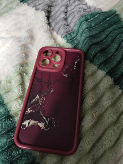 The image shows a close-up, high-angle view of a dark red phone case with a marble-like design in gold and black. The phone case is positioned diagonally on a plush, textured blanket. The blanket has wavy stripes of different colors, including off-white, grey, and dark green. The phone case has cutouts for a camera module on the top left, suggesting it's for a smartphone. The lighting is soft and even, with no harsh shadows, and the overall mood is calm and still. There is no visible text in the image. The setting appears to be indoors, possibly a home, given the presence of a blanket. The specific location of Vysokyi, Ukraine, is provided as context but cannot be visually inferred from the image itself.