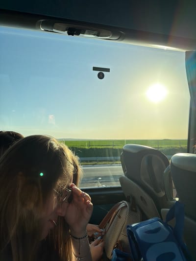 The image, rotated ninety degrees counterclockwise, depicts a person sitting inside a vehicle, likely a bus, in Градина, Bulgaria. The sun shines brightly through the window, illuminating a field of green to the right. A black object hangs from the window near the top left corner. The person's face is partially obscured by their hair. They have their hands raised near their face, and wear bracelets on their wrist. A grey bag and a blue bag are visible above the person's head, partially obstructing the view through the window. The interior of the vehicle appears to be grey.