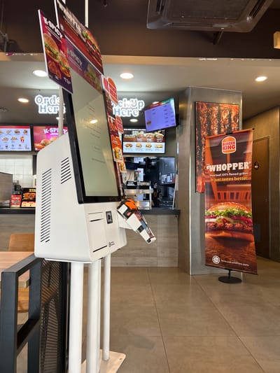 This is a photograph taken inside a Burger King restaurant, likely in Subang Jaya City Council, Malaysia, given the presence of a HALAL certification logo. The scene focuses on a self-order kiosk to the left, with its screen displaying a confirmation message stating "Payment Successful" and offering options to return to the main screen or home screen. The kiosk has a card reader and a receipt printer.
Behind the kiosk, a counter area is visible with digital menu boards displaying food items and prices, and what appears to be a pickup area labeled "Pick Up Here". A Burger King employee wearing a face mask and uniform is partially visible behind the counter.
To the right of the counter, a large vertical banner advertising "The Whopper" with its slogan "Real beef. 100% flamed-grilled. It just tastes better." is prominently displayed. The flooring is tiled, and the lighting suggests an indoor environment. Signs indicating "Order Here" are also visible. The overall impression is of a typical fast-food restaurant setting.