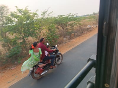 Captured from within a moving vehicle, the image shows a man driving a motorcycle with a woman riding on the back and a small child sitting in front of him on the seat of the bike. The man is wearing a red shirt and jeans, while the woman has on a green sari. They are traveling along a paved road that is bordered by trees and shrubs on one side and open fields on the other. The photo was taken in Kothacheruvu, India.